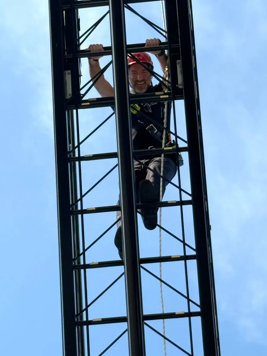 Close view of the EMS Medical Director in a helmet and safety harness climbing high on a fire department aerial ladder against a blue sky.