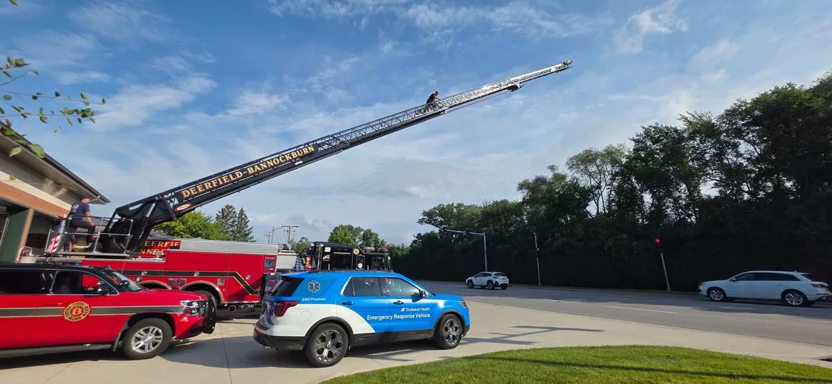 An EMS physician climbs the extended aerial ladder of a Deerfield-Bannockburn fire truck during training, with the Highland Park EMS System physician response vehicle parked below.